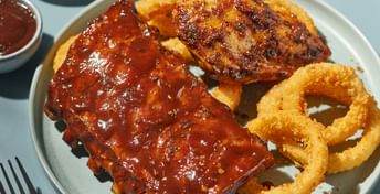 Plate of barbecue ribs and onion rings with coleslaw, sauce, and a fork on a light blue surface with a red napkin.