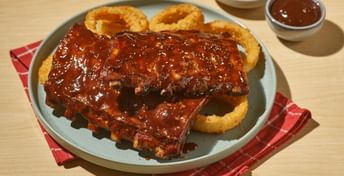 Glazed barbecue ribs with onion rings on a white plate, served with coleslaw and sauce on a wooden surface with red napkin.