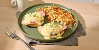 Eggs Benedict breakfast plate with hash browns, served on a green ceramic dish with coffee and fork on a wooden table with white subway tile backsplash