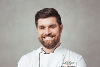 Professional headshot of a chef wearing a white chef's coat with White Spot restaurant logo and name "Dillon Penman" embroidered on it, smiling at camera against neutral background