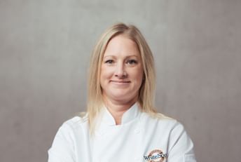 Professional portrait of a chef in a white chef's coat with arms crossed, smiling at the camera against a neutral gray background.