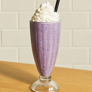 Purple berry smoothie milkshake topped with whipped cream and a black straw, served in a tall glass on a wooden surface against a cream subway tile background.