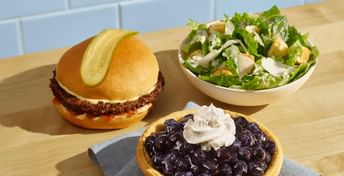 A Legendary Burger with a pickle slice on top, side of Caesar salad, and a BC Blueberry Pie, all on a wooden counter with blue subway tile backdrop.