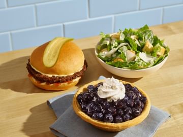A Legendary Burger with a pickle slice on top, side of Caesar salad, and a BC Blueberry Pie, all on a wooden counter with blue subway tile backdrop.
