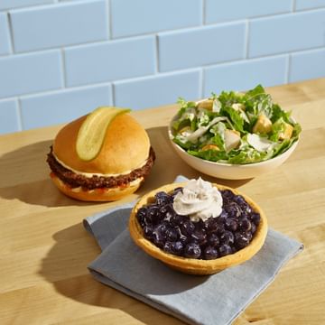 A Legendary Burger with a pickle slice on top, side of Caesar salad, and a BC Blueberry Pie, all on a wooden counter with blue subway tile backdrop.