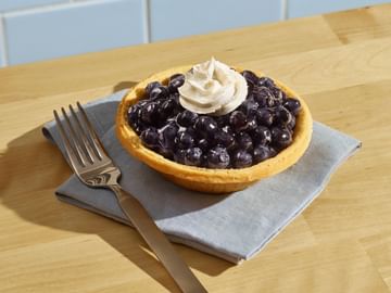 Berry pie with whipped cream topping on a gray napkin, accompanied by a fork, displayed on a wooden table with blue subway tile backdrop.