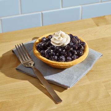 Berry pie with whipped cream topping on a gray napkin, accompanied by a fork, displayed on a wooden table with blue subway tile backdrop.