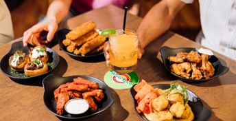 People sharing appetizers and drinks at a dining table, including wings, fried items, and a yellow cocktail with lime garnish on black plates.