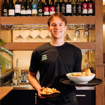 Smiling White Spot server in black uniform holding plates of food and fries in front of a wine display with bottles and hanging glasses in a restaurant setting.