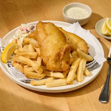 Fish and chips meal on a wooden table with coleslaw, lemon wedges, tartar sauce, and a fork on the side