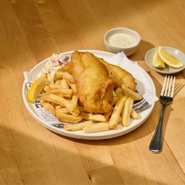 Classic fish and chips meal served on a white plate with coleslaw, golden fried fish fillet, thick-cut fries, lemon wedges, tartar sauce, and a fork on a wooden table.