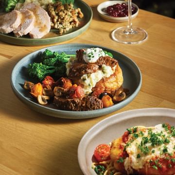 Elegant dinner table with three plated dishes featuring sliced poultry, roasted vegetables, mashed potatoes, and broccoli, accompanied by a glass of red wine and small bowl of cranberry sauce, set against a festive backdrop of twinkling warm lights and evergreen garland.