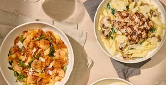 Three plates of pasta dishes on a light wood table with napkins and forks, featuring shrimp scampi, tagliatelle bolognese, and creamy mushroom pasta garnished with herbs and cheese.