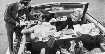 Vintage black and white photograph of a convertible car in a White Spot parking lot with a uniformed server delivering food to smiling passengers seated inside the vehicle.