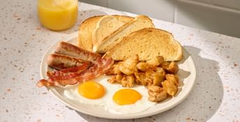 Full breakfast plate with two fried eggs, bacon strips, sausages, and hash browns, served with toasted bread slices and orange juice on a speckled countertop.