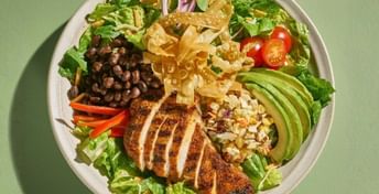 Overhead view of a colorful salad bowl with sliced chicken breast, black beans, avocado, crispy tortilla strips, corn, red peppers, onions, and lettuce on a green background.