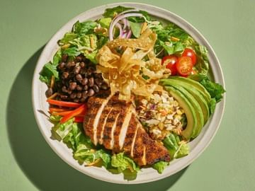 Overhead view of a colorful salad bowl with sliced chicken breast, black beans, avocado, crispy tortilla strips, corn, red peppers, onions, and lettuce on a green background.