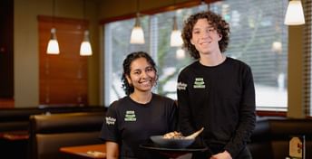 Two people wearing White Spot branded black shirts stand in a modern restaurant setting, smiling at the camera while holding a bowl of food.