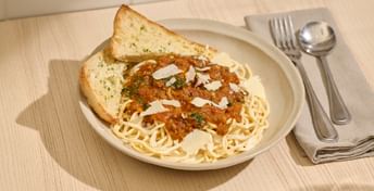 Spaghetti bolognese served on a white plate with garlic bread, topped with shaved cheese and fresh herbs, accompanied by fork and spoon on a light wooden table.