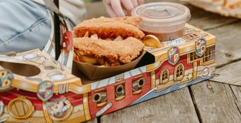 Child's hand reaching for fried chicken in a decorative cardboard container on a wooden table, with sauce cup and other food visible nearby.
