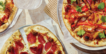 Overhead view of four freshly baked pizzas on white plates with various toppings including pepperoni, vegetables, and melted cheese on a checkered tablecloth