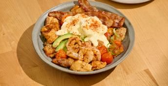 Breakfast dish with scrambled eggs, bacon, sausage, shrimp, potatoes, tomatoes, avocado slices, and paprika, served on a gray plate with toast on the side.