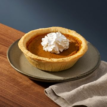 Pumpkin pie in a fluted pastry shell topped with whipped cream, served on a gray ceramic plate with a beige napkin on a wooden surface against a dark blue background