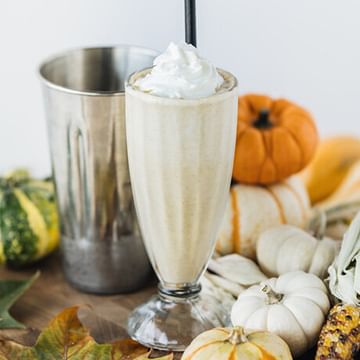 Pumpkin spice milkshake topped with whipped cream in a tall glass, surrounded by a metal shaker, fresh pumpkins, gourds, and autumn leaves on a wooden surface
