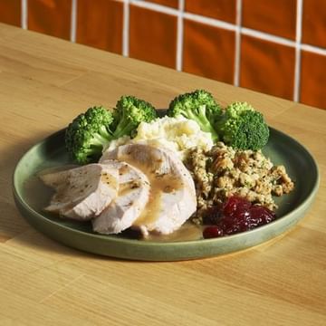 Plate with sliced turkey breast, steamed broccoli, mashed potatoes, ground meat mixture, and cranberry sauce on a wooden table with brick wall background