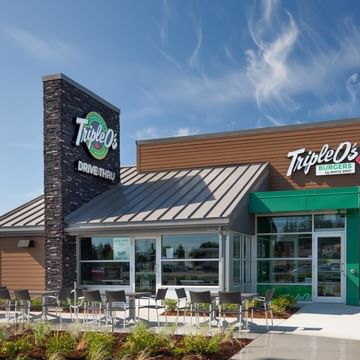 Triple O's Drive-Thru burger restaurant with modern wood and brick exterior, green entrance, tall sign, outdoor seating area, and tall evergreen trees under blue sky.