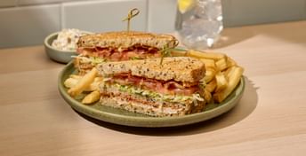 BLT sandwich on seeded bread with lettuce and bacon, served with French fries on a sage green plate, accompanied by a small bowl of sauce and a glass of lemon water.