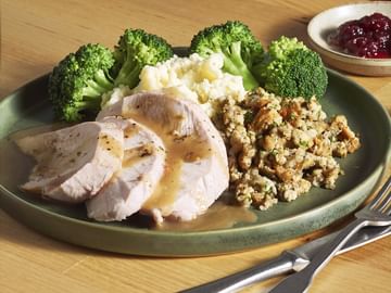Plate of sliced turkey breast with mashed potatoes, broccoli florets, and herb stuffing, served with gravy and a small bowl of cranberry sauce on a wooden table