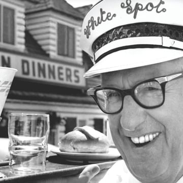 Black and white vintage photo of Nat Bailey smiling wearing glasses and a white chef's hat at a dining table with beverages, with a "Chicken Dinners" restaurant sign visible in the background.