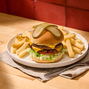 Cheeseburger with lettuce and pickles topped with sliced jalapeños, served with french fries on a white plate, set on a napkin with utensils, against a red background
