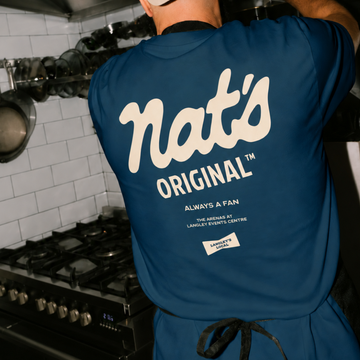 Person wearing navy blue "Nat's Original" sweatshirt standing in a kitchen with professional cooking equipment and white subway tiles visible in the background.