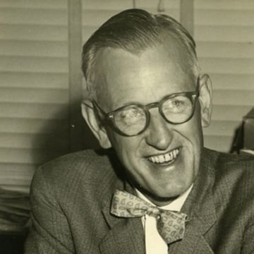 Black and white photograph of a smiling person wearing round glasses, a gray suit jacket, and a patterned bow tie, positioned against a brick wall background.