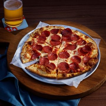 Pepperoni pizza on a white plate with one slice removed, served on a wooden table alongside a glass of beer with a coaster